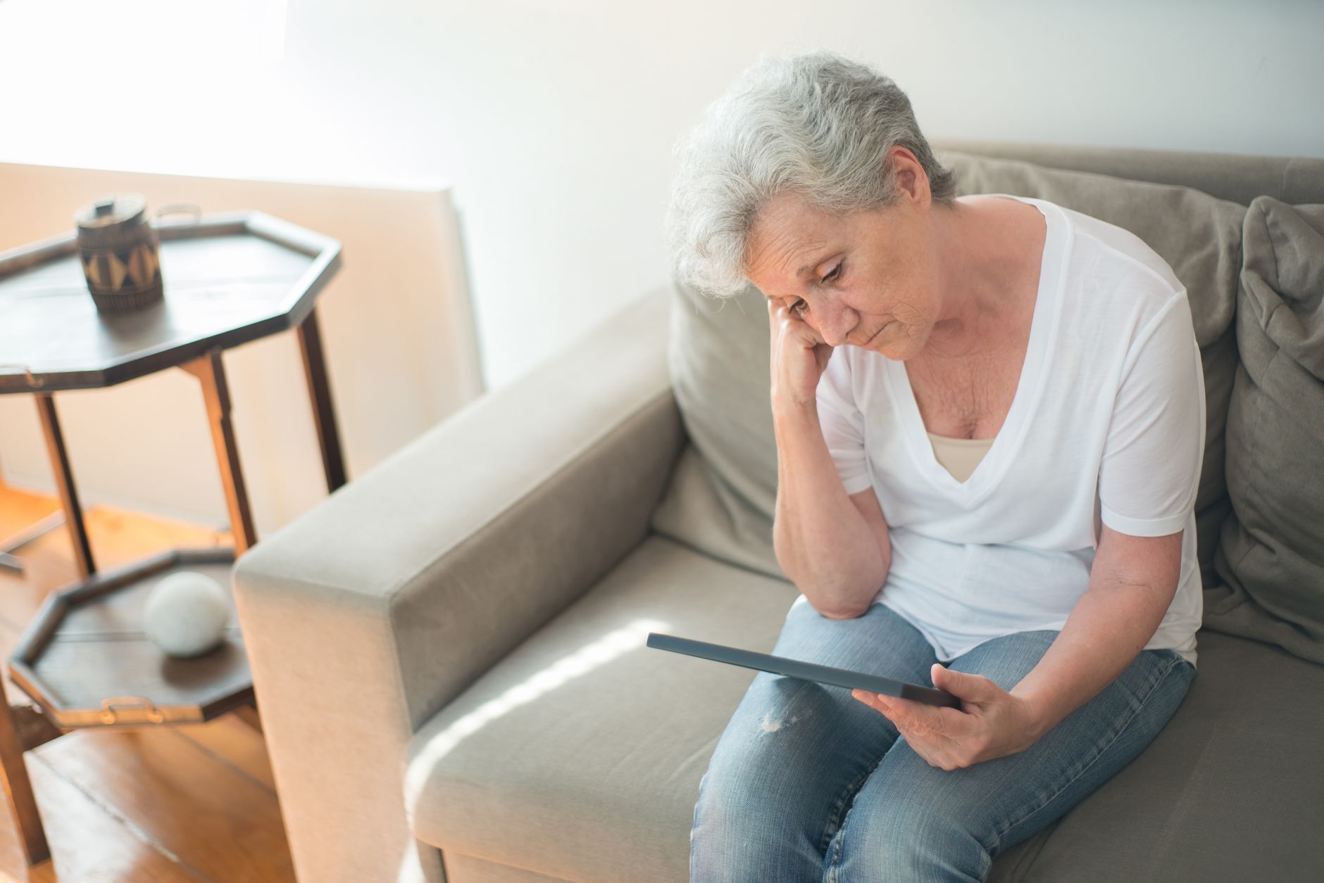 Woman sitting thoughtfully on a sofa in a bright living room, holding a tablet