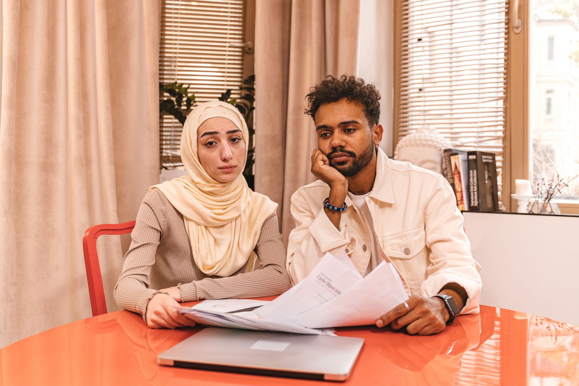 A couple sits at a glossy table in a warm living room with a pile of paperwork and a closed laptop in front of them.