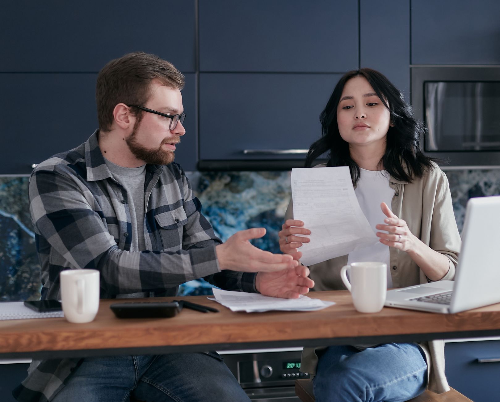 A man and a woman at a kitchen counter talk over a printed document, with two mugs, a laptop and a calculator in front of them.