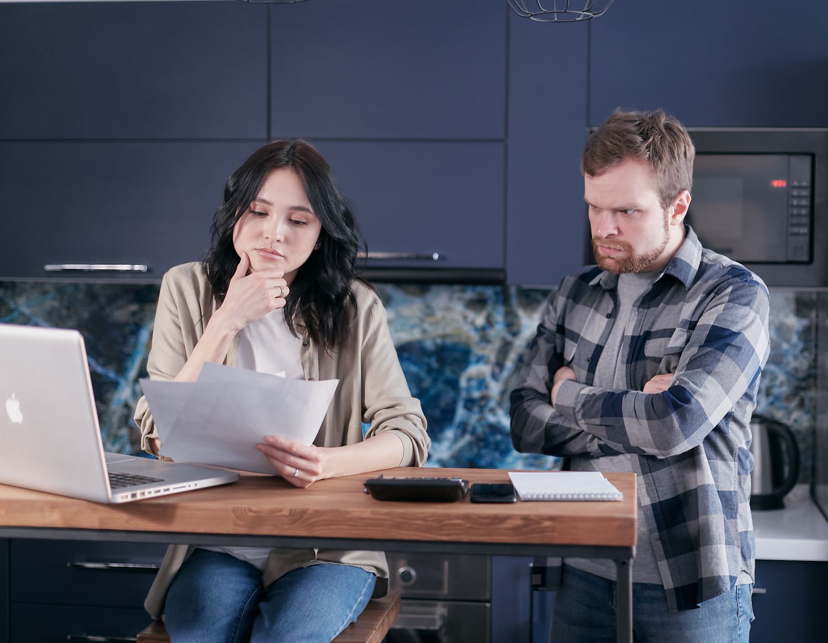 A woman holds a sheet of paper over a kitchen counter while a man stands beside her, with a laptop, calculator and notepad on the counter.